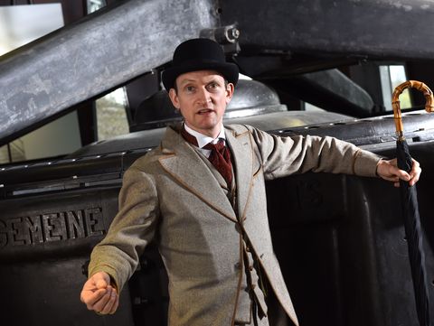 Eine Person in historischer Kleidung, die einen Anzug und einen Hut trägt, steht vor einer großen industriellen Maschine und hält einen Regenschirm. Das Bild thematisiert Emil Rathenau, einen bedeutenden deutschen Industriellen und Gründer der Allgemeinen Elektricitäts-Gesellschaft (AEG)