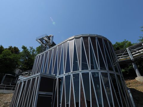 Ausstellungspavillon "Kraftwerk 1898" mit einer Glas- und Metallfassade, das Teil der Infrastruktur von naturenergie ist. Im Hintergrund sind grüne Bäume und ein klarer blauer Himmel zu sehen.