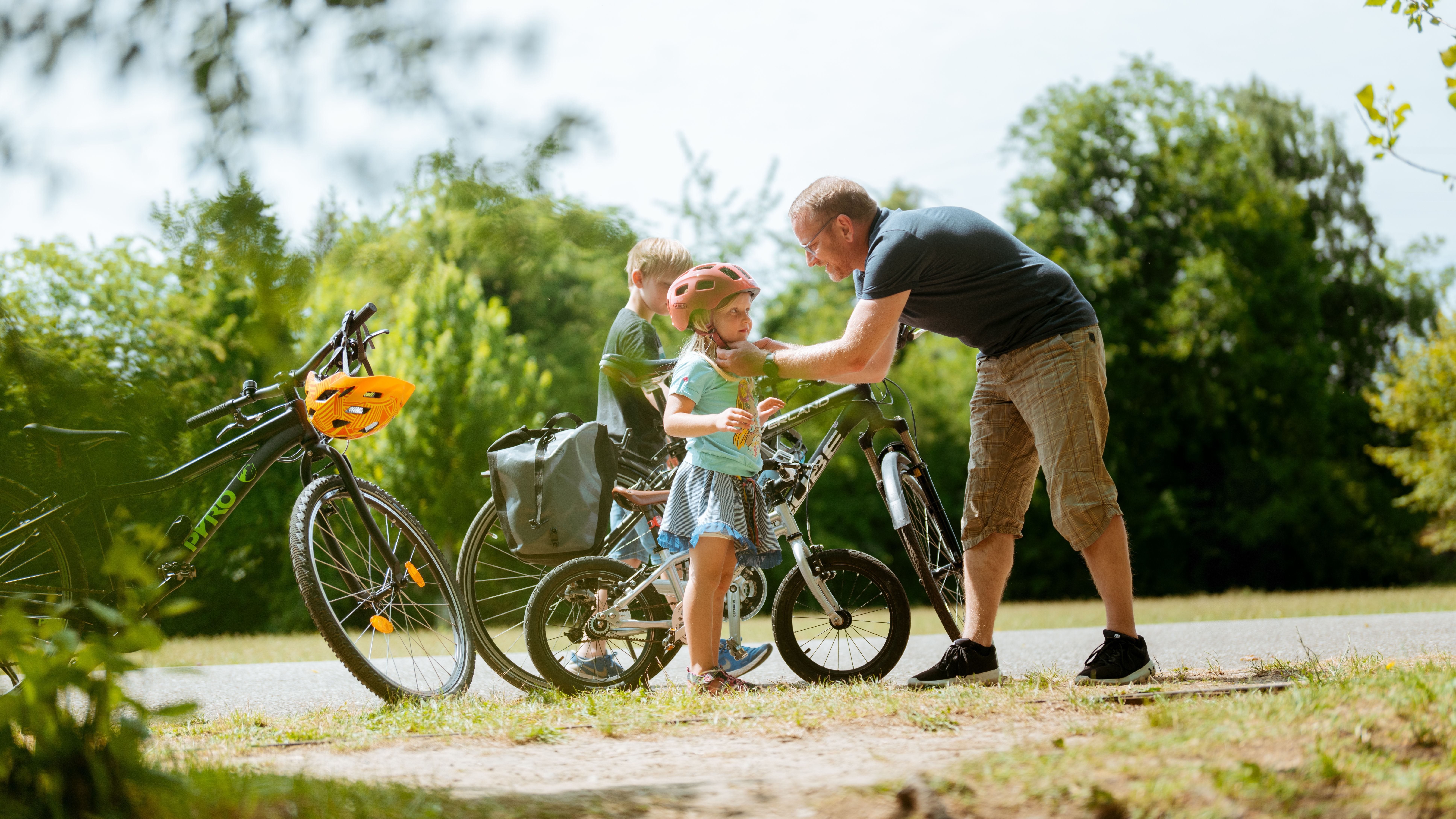 Ein Mann, wahrscheinlich der Papa, hilft einem kleinen Mädchen, das einen Fahrradhelm trägt, während ein Junge daneben steht. Im Hintergrund sind Fahrräder und grüne Bäume zu sehen.