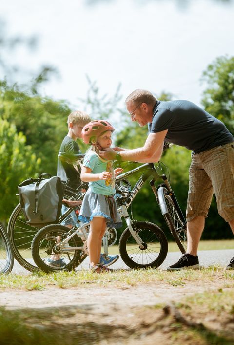 Ein Mann, wahrscheinlich der Papa, hilft einem kleinen Mädchen, das einen Fahrradhelm trägt, während ein Junge daneben steht. Im Hintergrund sind Fahrräder und grüne Bäume zu sehen.