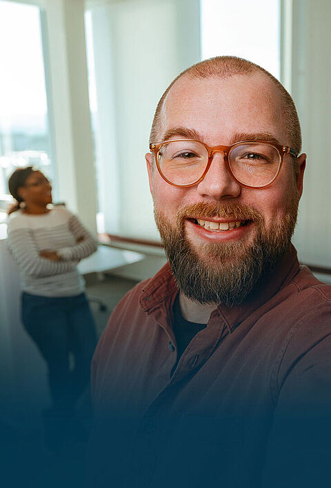 Ein lächelnder Mann mit Brille macht ein Selfie im Büro. Im Hintergrund sieht man drei seiner Kollegen bei der Arbeit.