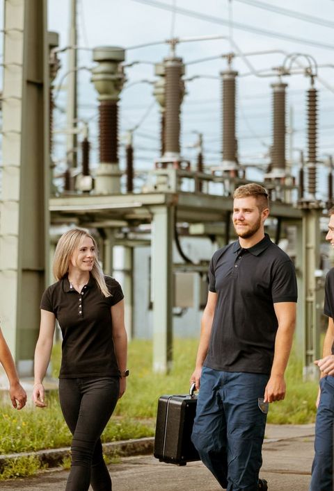 Vier Personen in schwarzen Poloshirts gehen durch eine industrielle Umgebung mit vielen Stromleitungen und Transformatoren. Sie tragen Werkzeuge und Ausrüstung.
