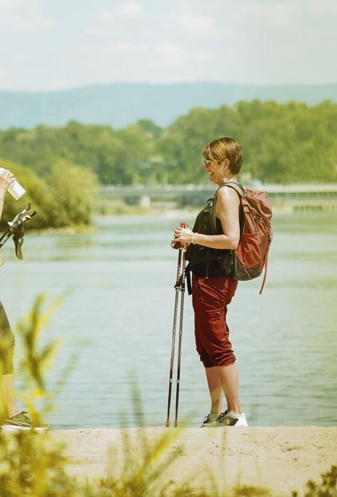 Zwei Damen mit Wanderstöcken und Freizeitkleidung machen eine Pause vor dem Rhein, im Hintergrund das Wasserkraftwerk von Rheinfelden.