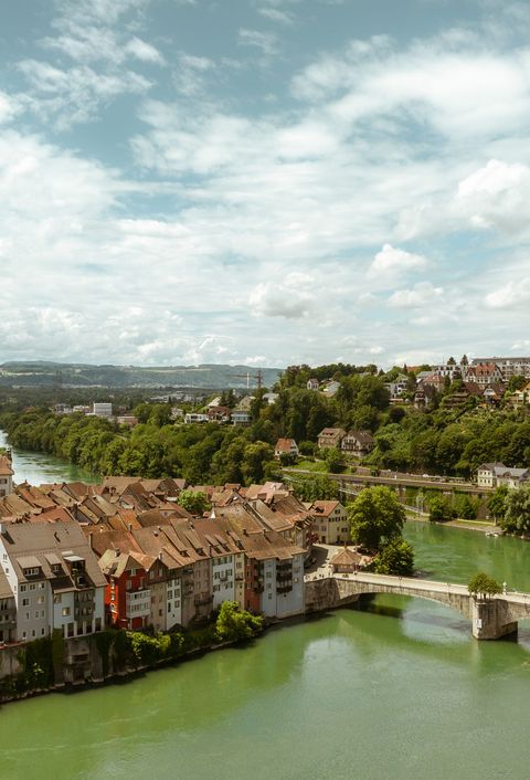 Panoramablick auf eine malerische Stadt mit einer Kirche, traditionellen Häusern und einer Brücke über einen Fluss, umgeben von grünen Hügeln und bewölktem Himmel.