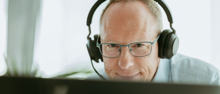 Eine Person sitzt an einem Schreibtisch und trägt große Kopfhörer. Im Hintergrund sind verschwommene Büromöbel und Fenster zu sehen.