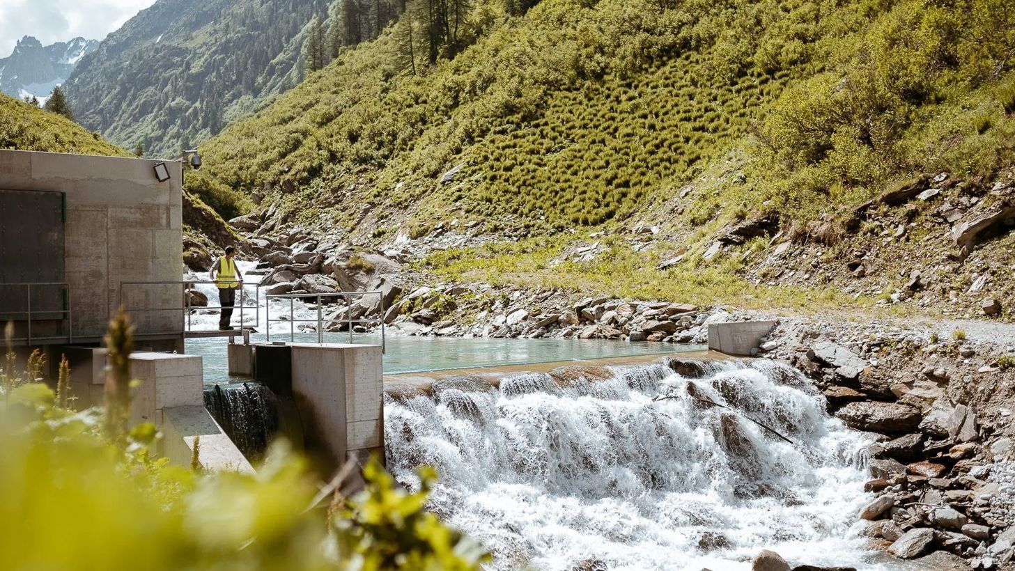 Ein Mitarbeiter von naturenergie in einer gelben Sicherheitsweste steht auf einer Plattform neben einem kleinen Wasserkraftwerk in einer bergigen Landschaft. Wasser fließt über eine Staumauer, umgeben von grüner Vegetation und felsigem Gelände
