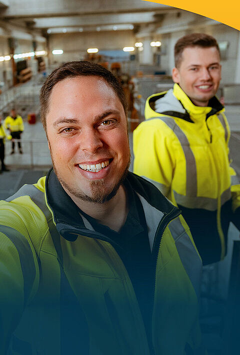 Ein lächelnder Mann macht ein Selfie in einer Maschinenhalle. Im Hintergrund sieht man mehrerer seiner Kollegen in Sicherheitswesten.