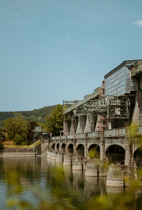 Ein Wasserkraftwerk von naturenergie, das sich über einen Fluss erstreckt. Die Struktur des Kraftwerks besteht aus mehreren Steinpfeilern und Metallbrücken. Im Hintergrund sind grüne Bäume und Hügel zu sehen