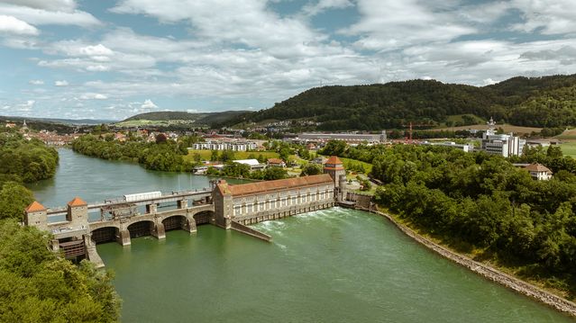 Ein Wasserkraftwerk von naturenergie in Laufenburg, das sich über einen Fluss erstreckt. Umgeben von grünen Hügeln und einer Stadtlandschaft im Hintergrund unter einem bewölkten Himmel.