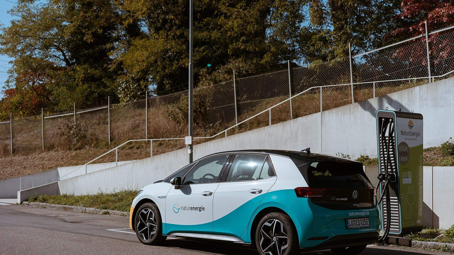 Elektroauto mit naturenergie-Logo steht an einer Ladestation und wird aufgeladen. Die Ladestation ist ebenfalls mit dem naturenergie-Logo versehen. Im Hintergrund sind eine Straße, ein Zaun und Bäume zu sehen.