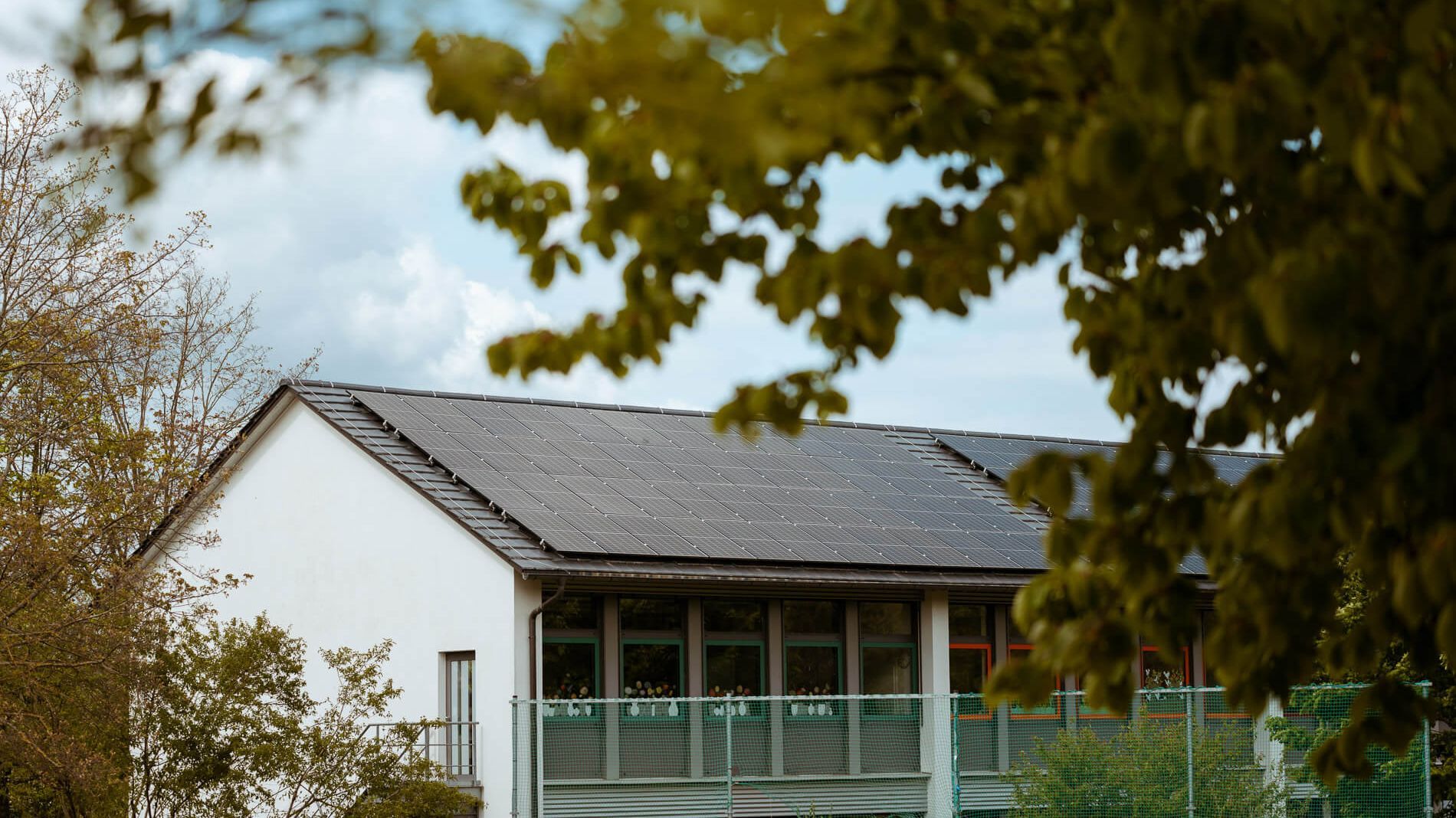 Ein Gebäude mit Solarpaneelen auf dem Dach, umgeben von grünen Bäumen. Die Solarpaneele sind Teil der nachhaltigen Energieerzeugung von naturenergie