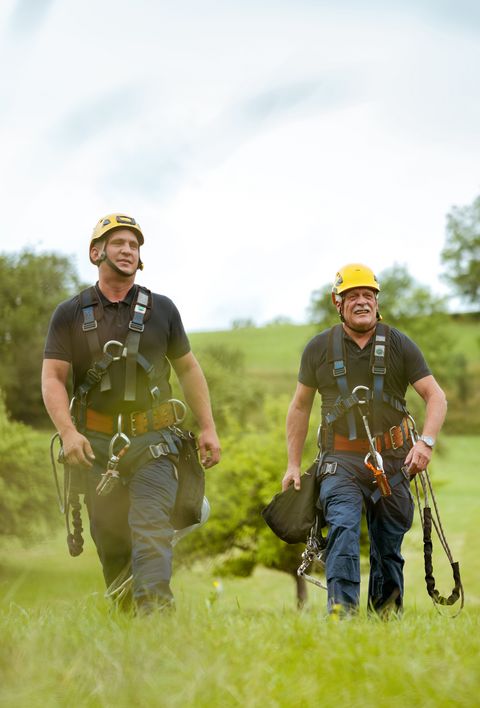 Zwei Männer in Arbeitskleidung und Sicherheitsausrüstung gehen über eine grüne Wiese. Im Hintergrund sind Bäume und eine hügelige Landschaft zu sehen.