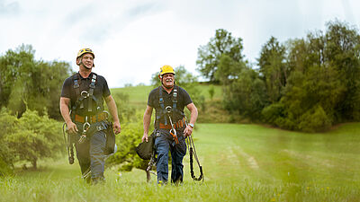 Zwei Männer in Arbeitskleidung und Sicherheitsausrüstung gehen über eine grüne Wiese. Im Hintergrund sind Bäume und eine hügelige Landschaft zu sehen.