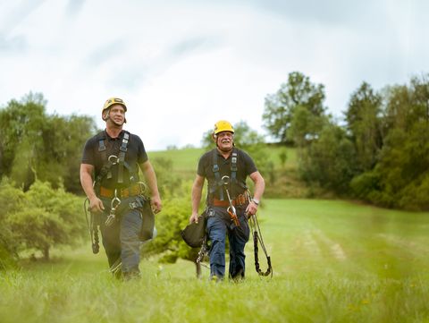 Zwei Männer in Arbeitskleidung und Sicherheitsausrüstung gehen über eine grüne Wiese. Im Hintergrund sind Bäume und eine hügelige Landschaft zu sehen.