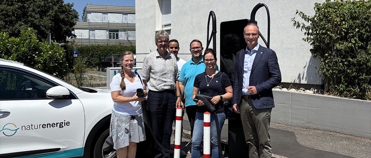 Fünf Personen stehen an einer Ladestation für Elektrofahrzeuge vor einem hellen Gebäude im Außenbereich. Im Vordergrund ist links ein weißes Elektroauto mit dem Logo „naturenergie“ zu sehen. Die Personen stehen neben der Ladesäule und halten teilweise Ladekabel in den Händen. Die Ladesäule befindet sich zwischen dem Fahrzeug und der Gruppe. Im Hintergrund sind weitere Gebäude, Bäume, Sträucher und ein Parkplatz erkennbar. Die Aufnahme entstand bei sonnigem Wetter und zeigt eine Situation rund um das Laden eines Elektrofahrzeugs.