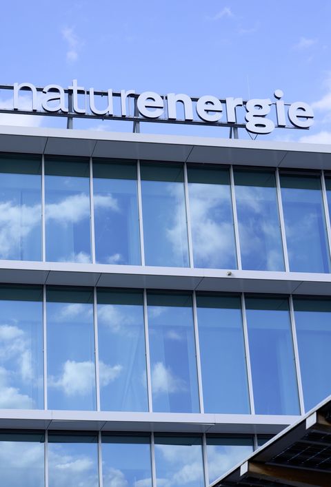Modernes Bürogebäude der Firma Naturenergie mit großen Glasfenstern und reflektierendem Himmel. Ein Baum steht im Vordergrund.