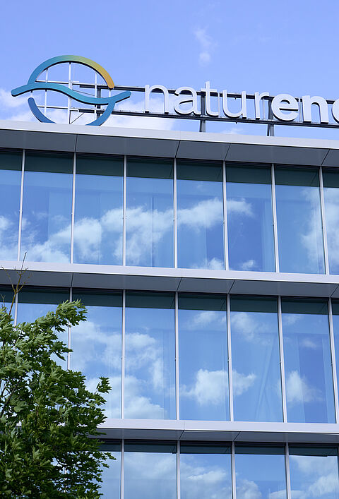 Modernes Bürogebäude der Firma Naturenergie mit großen Glasfenstern und reflektierendem Himmel. Ein Baum steht im Vordergrund.