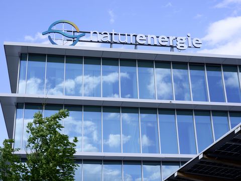 Modernes Bürogebäude der Firma Naturenergie mit großen Glasfenstern und reflektierendem Himmel. Ein Baum steht im Vordergrund.