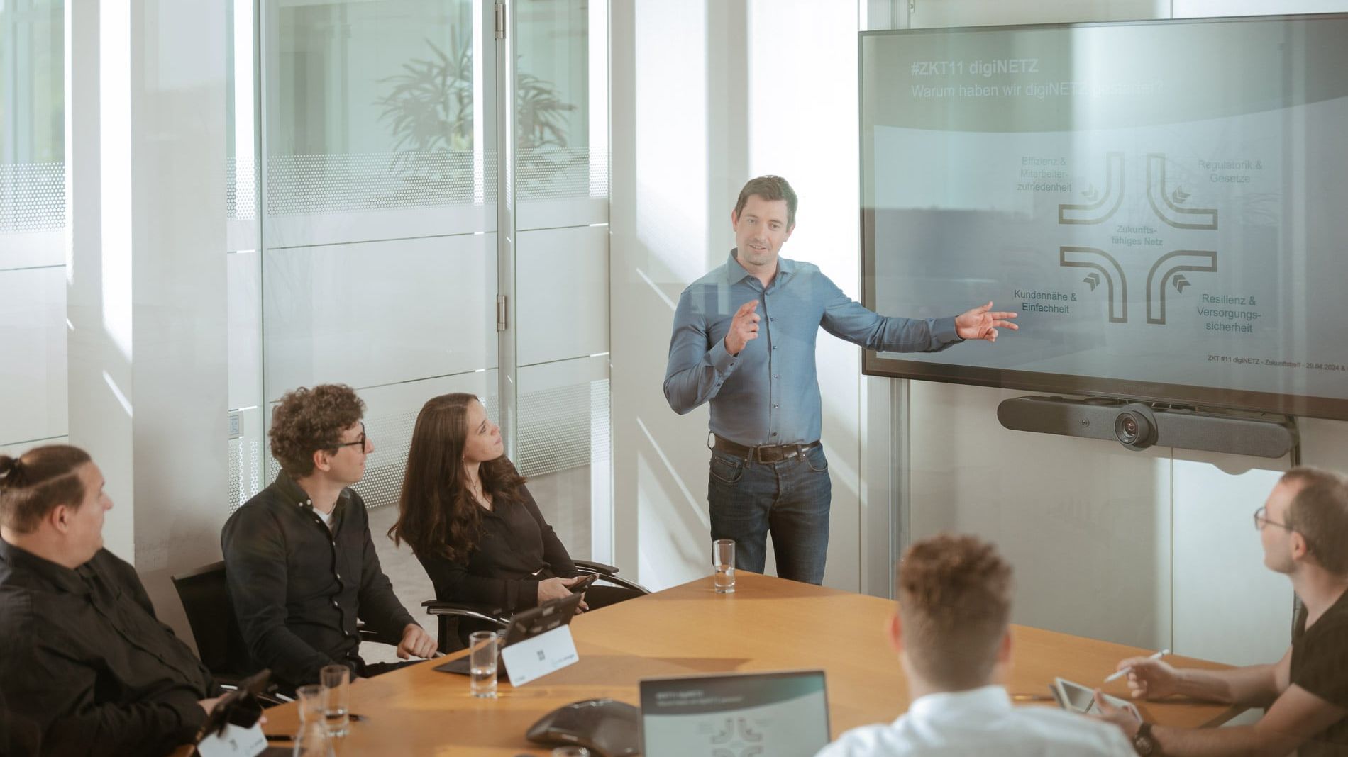 Eine Person steht vor einem Bildschirm und hält eine Präsentation. Mehrere Personen sitzen an einem Konferenztisch und hören zu. Der Raum ist modern und hell.