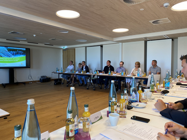 Gruppe von Menschen, die an einem großen Konferenztisch in einem modernen Raum sitzen. Auf dem Tisch stehen zahlreiche Flaschen und Gläser. Im Hintergrund ist ein Bildschirm mit einer Präsentation zu sehen.