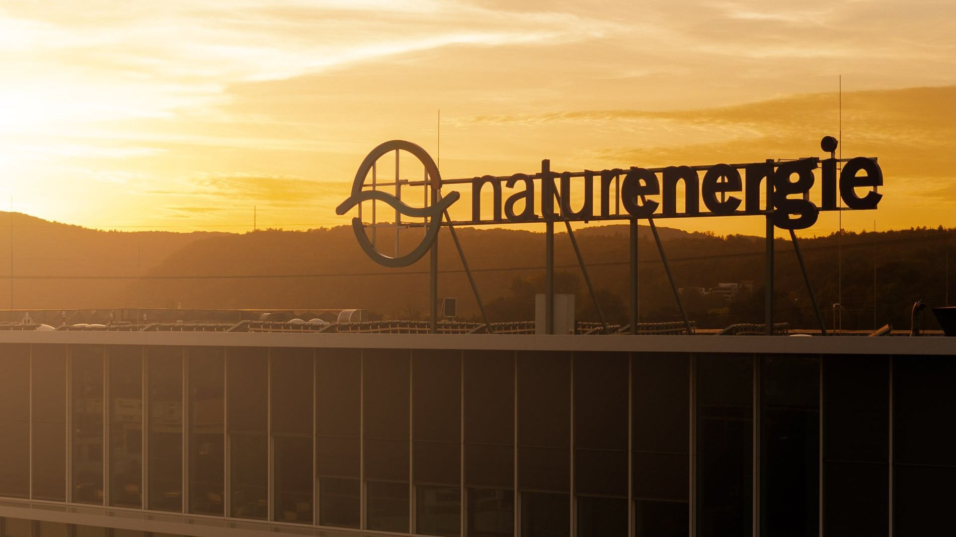 naturenergie-Logo auf einem Gebäude bei Sonnenuntergang. Der Himmel ist orange und gelb gefärbt, und im Hintergrund sind Hügel und Bäume zu sehen.
