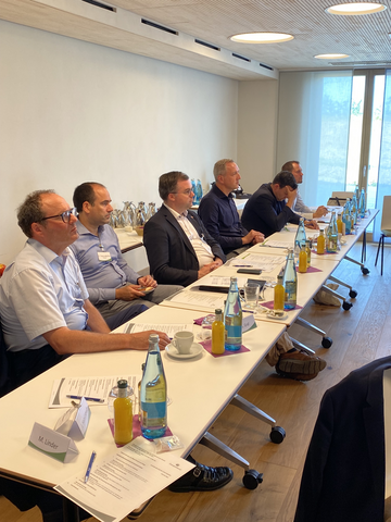Gruppe von Menschen, die an einem großen Konferenztisch in einem modernen Raum sitzen. Auf dem Tisch stehen zahlreiche Flaschen und Gläser. Im Hintergrund ist ein Bildschirm mit einer Präsentation zu sehen.