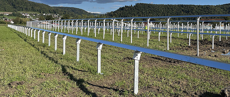 Reihen metallischer Unterkonstruktionen auf einer Wiese für einen entstehenden Freiflächen-Solarpark, mit Hügeln und Dorf im Hintergrund.