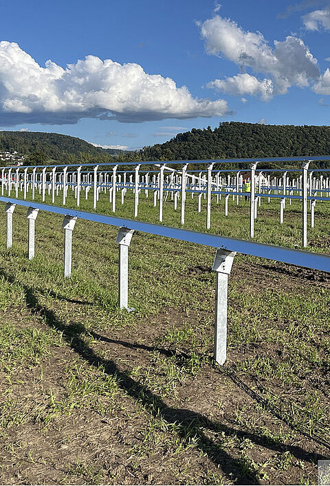 Reihen metallischer Unterkonstruktionen auf einer Wiese für einen entstehenden Freiflächen-Solarpark, mit Hügeln und Dorf im Hintergrund.