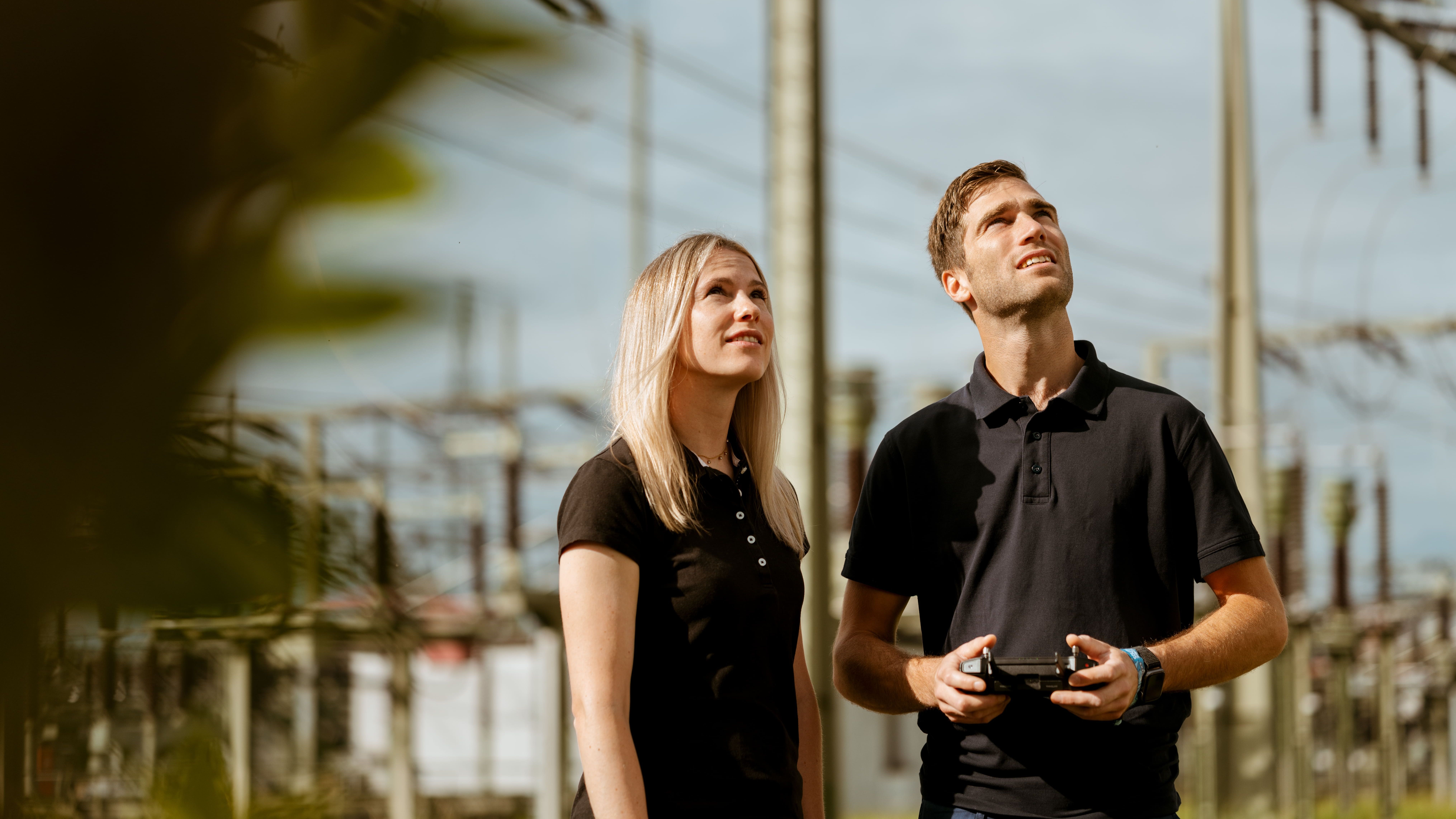 Zwei Personen in schwarzen Poloshirts stehen in einer industriellen Umgebung mit vielen Stromleitungen und Transformatoren. Eine Person hält eine Fernsteuerung in der Hand.