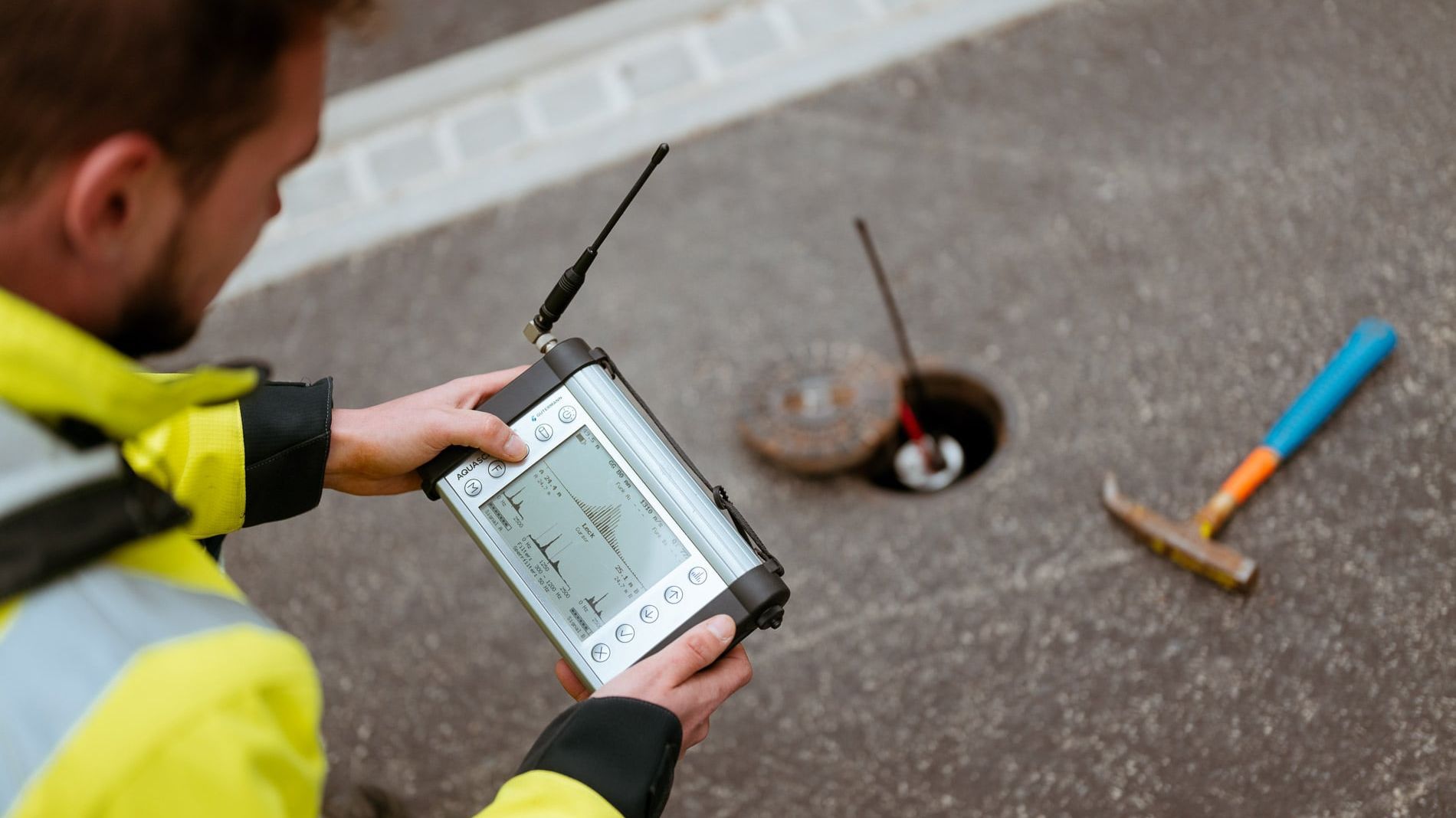 Eine Person in einer gelben Sicherheitsjacke bedient ein technisches Gerät mit einem Bildschirm und einer Antenne. Im Hintergrund ist ein offener Schacht auf einer Asphaltfläche zu sehen, neben dem ein Hammer liegt.