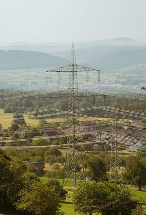 weite Landschaft mit grünen Feldern und Hügeln. Im Vordergrund sind Stromleitungen und ein Strommast zu sehen, die sich durch die Landschaft ziehen. Im Hintergrund sind Häuser und Bäume zu sehen