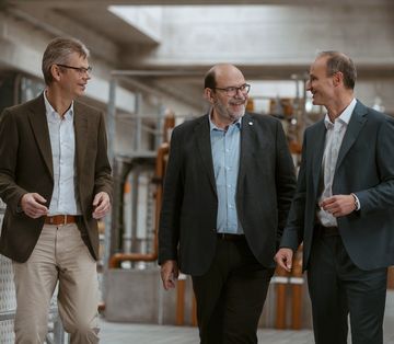 Die Geschäftsführung, bestehend aus Michel Schwery, Klaus Müller und Daniel Schölderle, im Gespräch in einem industriellen Umfeld. Sie tragen Business-Kleidung und befinden sich in einem technischen Gebäude.