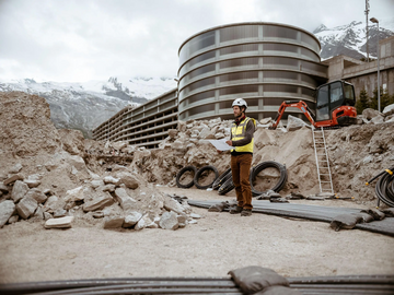 Ein Mitarbeiter von naturenergie in einer gelben Sicherheitsweste und einem weißen Helm steht auf einer Baustelle und betrachtet Baupläne. Im Hintergrund sind ein modernes Gebäude und schneebedeckte Berge zu sehen