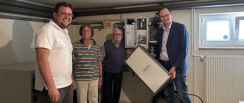 Vier Personen stehen in einem Technikraum vor einer geöffneten Heizungs- oder Energieanlage. Links steht eine Person in einem weißen Poloshirt, in der Mitte zwei ältere Personen, rechts eine Person in dunklem Sakko und Jeans. Die Person rechts hält ein rechteckiges technisches Bauteil mit angeschlossenem Kabel. Im Hintergrund sind Rohrleitungen, Steuerungstechnik und ein geöffneter Schaltschrank zu sehen. Rechts im Raum befinden sich ein Fenster und ein Heizkörper. Der Raum ist hell beleuchtet und wirkt wie ein Keller- oder Hausanschlussraum.