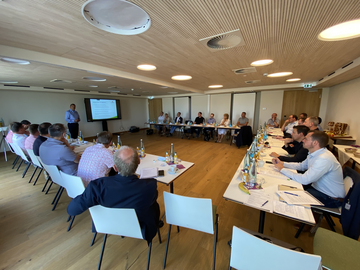 Gruppe von Menschen, die an einem großen Konferenztisch in einem modernen Raum sitzen. Auf dem Tisch stehen zahlreiche Flaschen und Gläser. Im Hintergrund ist ein Bildschirm mit einer Präsentation zu sehen.