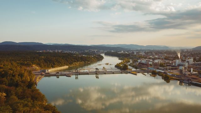 Ein Wasserkraftwerk von naturenergie in Rheinfelden, das sich über einen breiten Fluss erstreckt. Umgeben von einer Stadtlandschaft und grünen Hügeln im Hintergrund unter einem bewölkten Himmel bei Sonnenuntergang