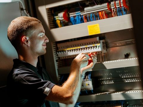 Ein Mann arbeitet an einem Schaltschrank mit vielen Kabeln und Schaltern. Sie verwendet ein Werkzeug, um an den elektrischen Komponenten zu arbeiten.