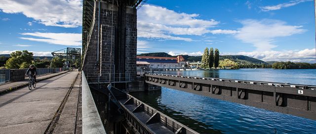 Ein Wasserkraftwerk von naturenergie in Whylen, das sich über einen Fluss erstreckt. Ein Radfahrer fährt auf einem Weg neben dem Kraftwerk. Im Hintergrund sind Gebäude und Bäume unter einem blauen Himmel mit Wolken zu sehen.