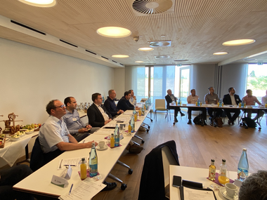 Gruppe von Menschen, die an einem großen Konferenztisch in einem modernen Raum sitzen. Auf dem Tisch stehen zahlreiche Flaschen und Gläser. Im Hintergrund ist ein Bildschirm mit einer Präsentation zu sehen.