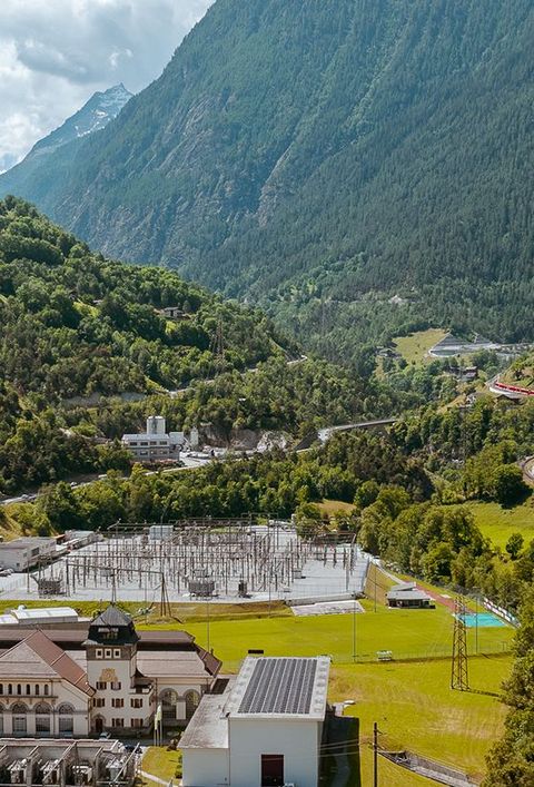 Gemeinde Ackersand, im Wallis, in einem Tal, umgeben von hohen, bewaldeten Bergen. Im Vordergrund befindet sich eine industrielle Anlage mit mehreren Gebäuden und Stromleitungen. Eine Straße und eine Eisenbahnlinie schlängeln sich durch das Tal. Der Himmel ist blau mit einigen Wolken.