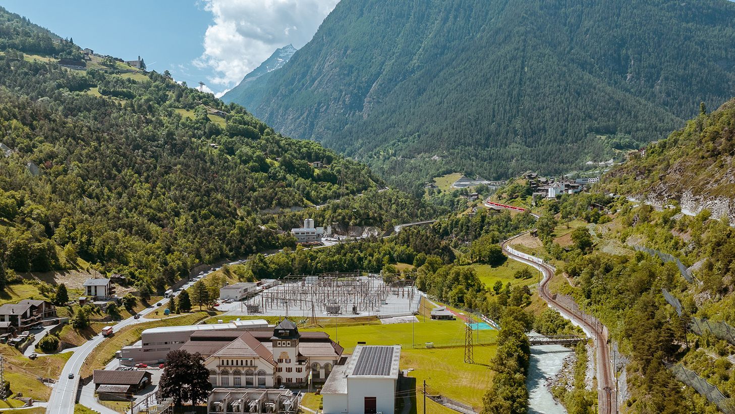 Gemeinde Ackersand, im Wallis, in einem Tal, umgeben von hohen, bewaldeten Bergen. Im Vordergrund befindet sich eine industrielle Anlage mit mehreren Gebäuden und Stromleitungen. Eine Straße und eine Eisenbahnlinie schlängeln sich durch das Tal. Der Himmel ist blau mit einigen Wolken.