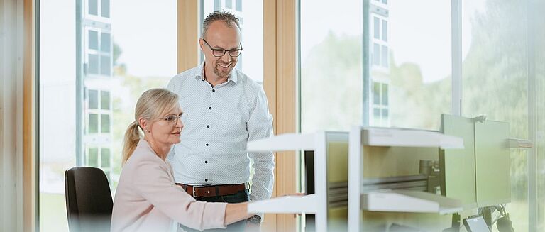Zwei Personen, Frau und Mann in einem Bürp am Schreibtisch unterhalten sich und zeige auf einen Bildschirm