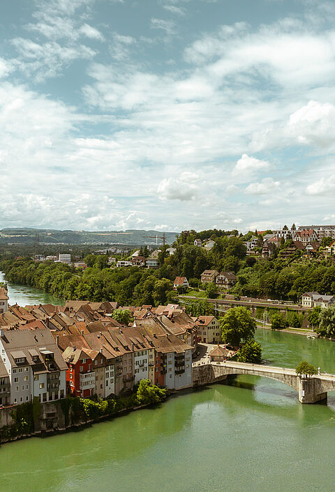 Panoramablick auf eine malerische Stadt mit einer Kirche, traditionellen Häusern und einer Brücke über einen Fluss, umgeben von grünen Hügeln und bewölktem Himmel.