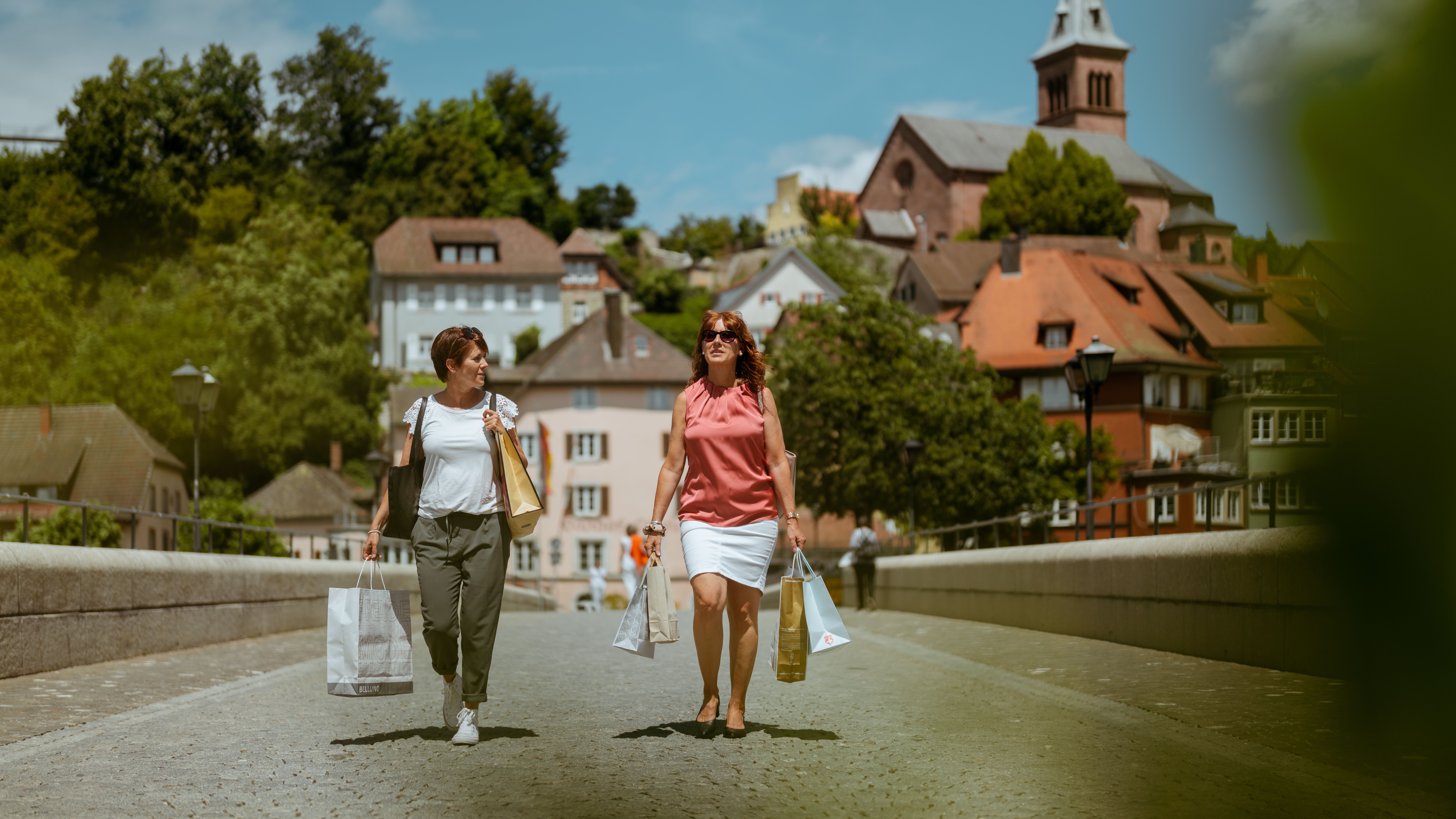 Zwei Frauen beim Einkaufen in einer malerischen Stadt mit Einkaufstaschen in der Hand. Im Hintergrund sind historische Gebäude und eine Kirche zu sehen.