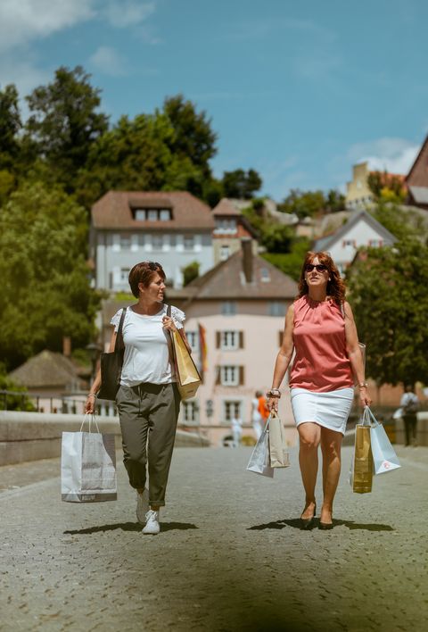 Zwei Frauen beim Einkaufen in einer malerischen Stadt mit Einkaufstaschen in der Hand. Im Hintergrund sind historische Gebäude und eine Kirche zu sehen.