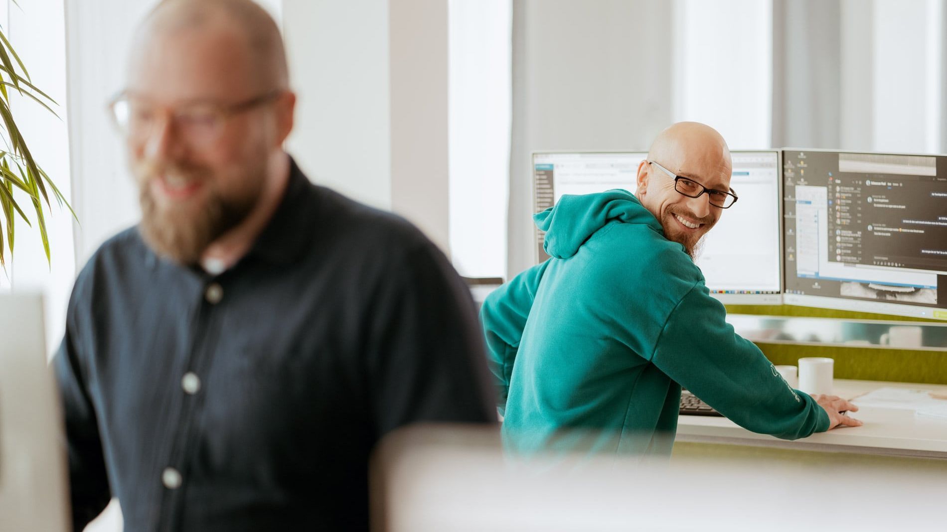 Zwei Männer arbeiten in einem Büro. Einer sitzt vor zwei Monitoren und trägt einen grünen Hoodie, während der andere im Vordergrund steht und eine schwarze Jacke trägt.