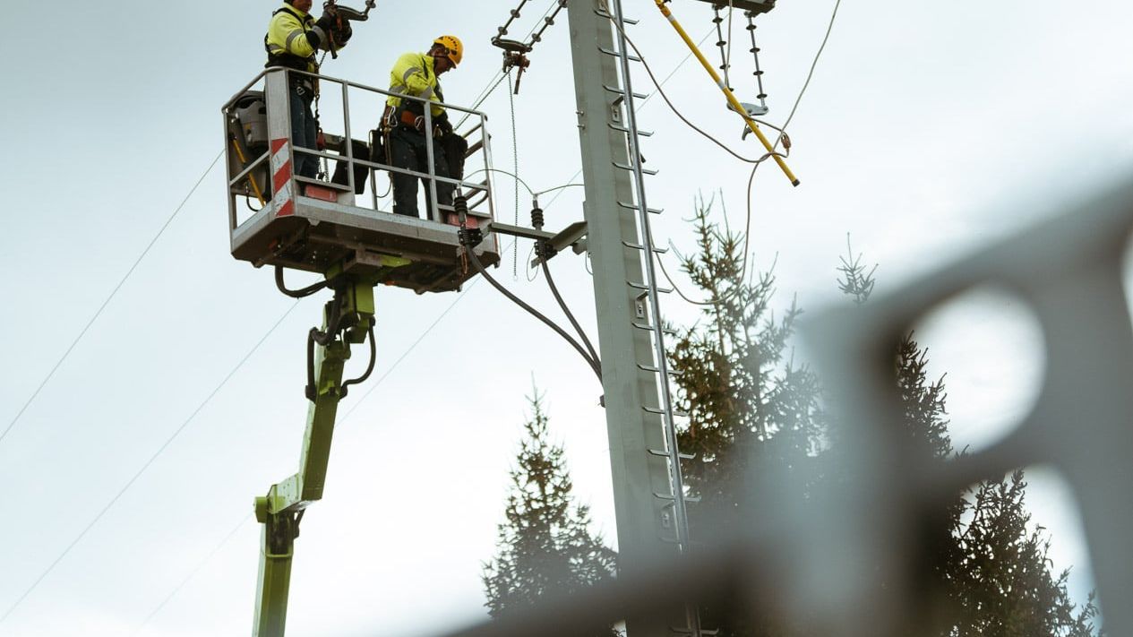 Zwei Arbeiter in gelben Sicherheitsjacken und Helmen stehen in einem Hebekorb und arbeiten an einer Stromleitung. Der Hebekorb ist an einem hohen Mast befestigt, und im Hintergrund sind Bäume und der Himmel zu sehen.
