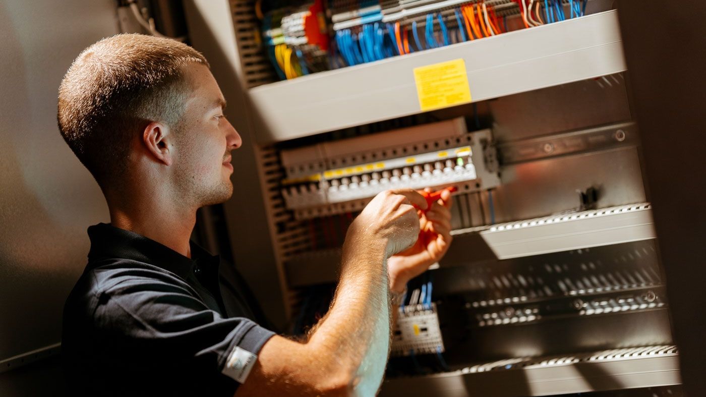 Eine Person arbeitet an einem Schaltschrank mit vielen Kabeln und Schaltern. Die Person trägt ein schwarzes Poloshirt und verwendet ein Werkzeug, um an den elektrischen Komponenten zu arbeiten.