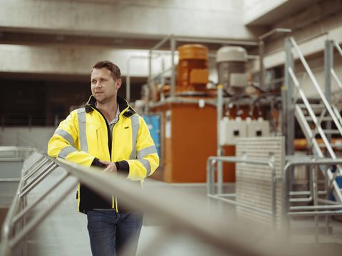 Ein Mann in einer gelben Sicherheitsjacke steht in einer industriellen Umgebung. Im Hintergrund sind verschiedene Maschinen und Metallstrukturen zu sehen.