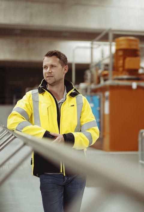Ein Mann in einer gelben Sicherheitsjacke steht in einer industriellen Umgebung. Im Hintergrund sind verschiedene Maschinen und Metallstrukturen zu sehen.